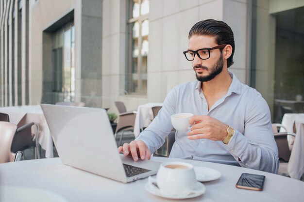 Geschäftsmann in Gläsern sitzt am weißen Tisch und arbeitet am Laptop. Er hält eine Tasse Kaffee in der Hand. Guy ist ernst und konzentriert. Telefon ist am Tisch.