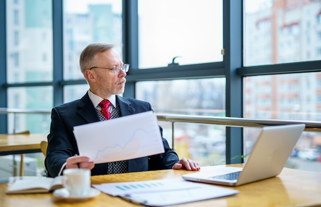 Geschäftsmann liest einen gedruckten Bericht oder ein Dokument Man sitzt am Tisch und arbeitet an Papierkram mit Laptop am Tisch Man schaut auf das Fenster mit Dokumenten in seinen Händen Business-Konzept