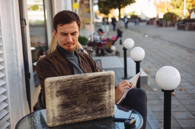 Geschäftsmann macht sich Notizen im Notizblock und benutzt Laptop, während er im Straßencafé am Tisch sitzt