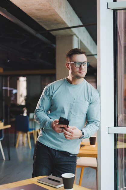 Geschäftsmann, Manager, der tief nachdenklich aus dem Fenster schaut und am Arbeitsplatz mit Telefon im Büro steht und über die Zukunft des Unternehmens nachdenkt und über eine Lösung für die Arbeitsaufgabe nachdenkt