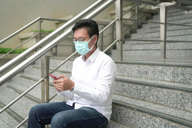 Geschäftsmann mit Gesichtsmaske sitzt auf der Treppe und benutzt und liest sein Telefon