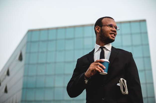 Geschäftsmann mit Kaffee zum Mitnehmen vor dem Wolkenkratzer. Schwarzer ethnischer Mann in Jacke und Krawatte