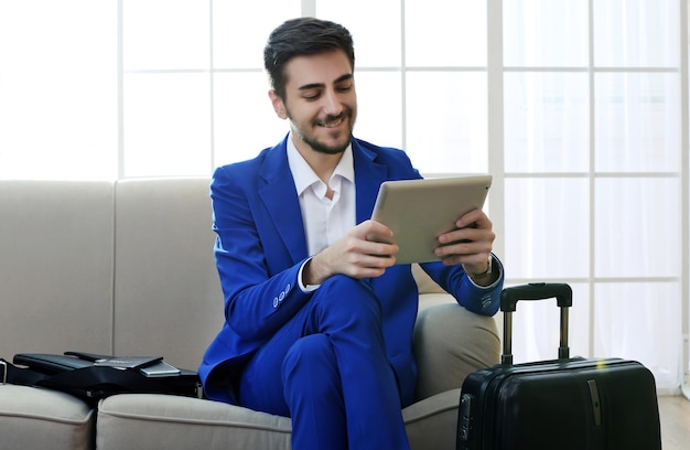 Geschäftsmann mit Koffer und Tablet sitzt auf dem Sofa in der Halle