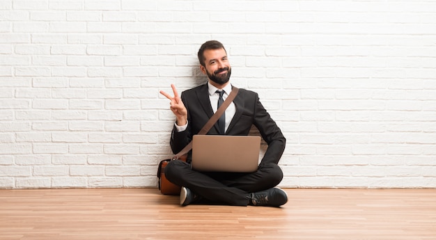 Geschäftsmann mit seinem Laptop, der auf dem Boden sitzt, lächelnd und Siegzeichen mit einem netten Gesicht zeigt