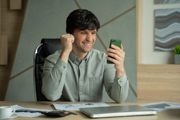 Geschäftsmann mit Smartphone, der im Büro sitzt und gute Nachrichten am Telefon hat