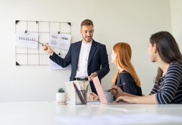 Geschäftsmann präsentiert und zeigt auf Verkaufsmarketing-Diagrammbericht auf Papier an Kolleginnen, während er sich im Zimmer im Büro trifft