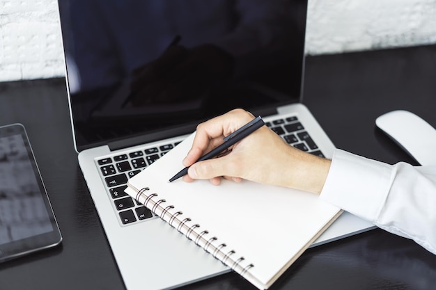 Geschäftsmann schreibt mit einem Stift in Tagebuch auf Laptop-Tastatur in einem sonnigen Büro, Geschäfts- und Bildungskonzept aus nächster Nähe