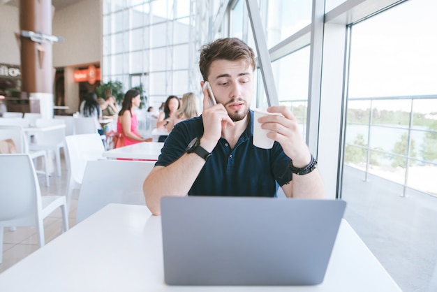 Geschäftsmann sitzt in einem Café mit einem Laptop und einem Glas Kaffee in der Hand, sagt ein Handy