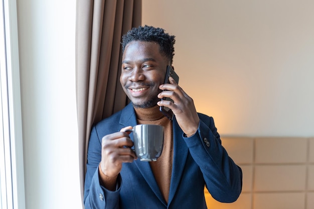 Geschäftsmann stand am Fenster im Hotelzimmer und trank Kaffee Nachdenklicher junger afrikanischer Geschäftsmann stand am Fenster mit einer Tasse Kaffee und schaute auf sein Handy