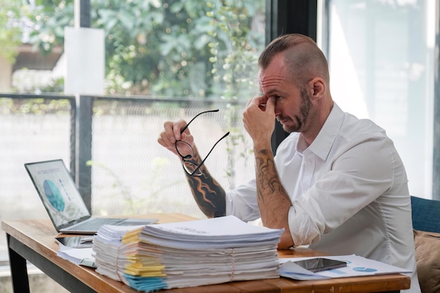 Geschäftsmann stresst Papierkram und Fristenkonzept. Gestresster Geschäftsmann mit Papieren und Diagrammen, der am Tisch im Büro sitzt