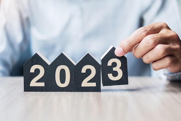 Geschäftsmannhände, die 2023 Frohes Neues Jahr mit Hausmodell auf Tischbüro halten Neues Haus Finanzielle Sachversicherung Immobiliensparen und Neujahrsvorsatzkonzepte