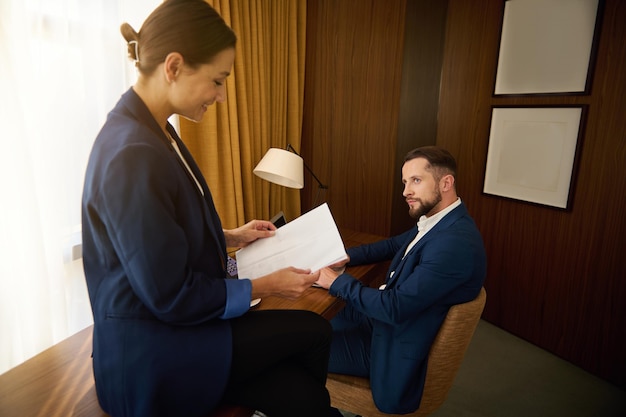 Geschäftspartner, die während der geschäftsreise im hotelzimmer zusammenarbeiten. gut aussehender mann, der aufmerksam zu seinem kollegen schaut, der dokumente liest, die auf dem tisch im hotelzimmer sitzen?