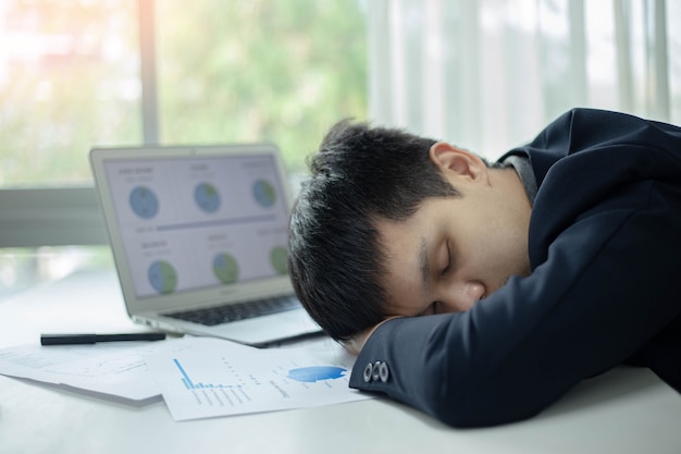 Geschäftspartner konzipieren einen männlichen jungen Geschäftsmann, der auf einem Schreibtisch mit einem Laptop und einem Dokument schläft, nachdem er lange Zeit gearbeitet hat.