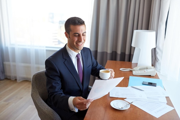 Geschäftsreise, Menschen und Papierkram Konzept - glücklicher lächelnder Geschäftsmann mit Papieren, der Kaffee im Hotelzimmer trinkt