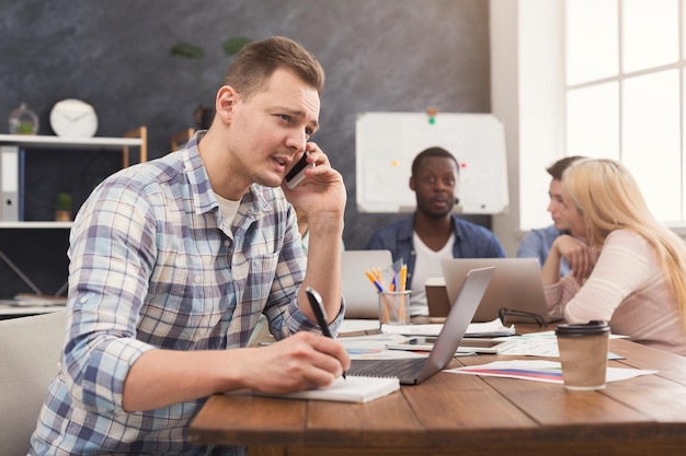 Geschäftsteam, das über Ergebnisse ihrer Arbeit spricht. Junger glücklicher Mann, der am Laptop arbeitet und telefoniert, während eine Gruppe von Geschäftsleuten über neue Marketingstrategien diskutiert, Platz kopieren