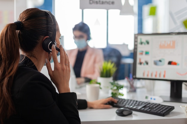 Geschäftsteam mit Headset, das in das Mikrofon spricht, Finanzstatistiken im neuen normalen Firmenbüro überprüft. Teamarbeiter, die Schutzmasken tragen, um eine Infektion mit dem Coronavirus zu verhindern.