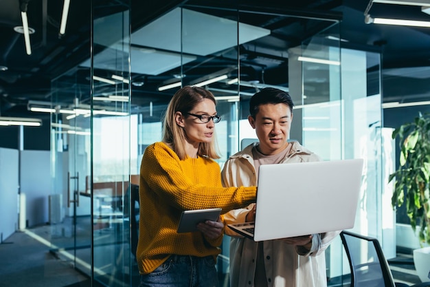 Geschäftstreffen zweier asiatischer und weiblicher Mitarbeiter, die über das Projekt diskutieren, das in einem modernen Büro arbeitet und auf einen Laptop-Bildschirm blickt, der Bearbeitungen und Projekterfolg bespricht