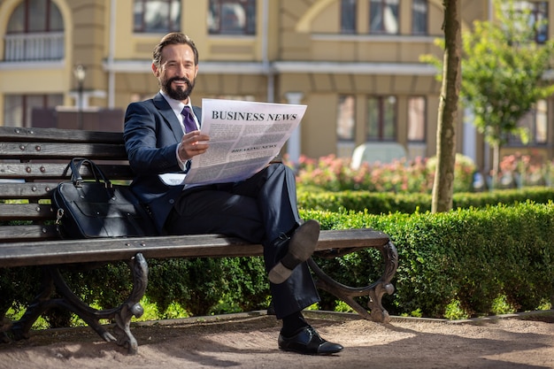 Geschäftstrends. Volle Länge eines lächelnden selbstbewussten Geschäftsmannes, der eine Zeitung liest, während er auf der Bank sitzt