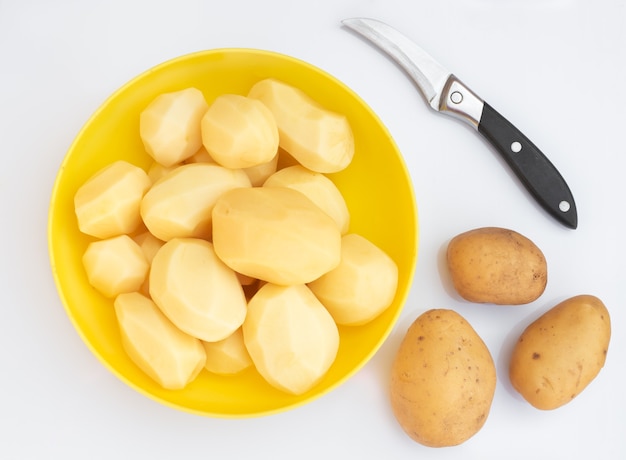 Foto geschälte kartoffeln in einer gelben tasse auf einer hellen oberfläche. in der nähe gibt es ein messer und kartoffelknollen. kochen.