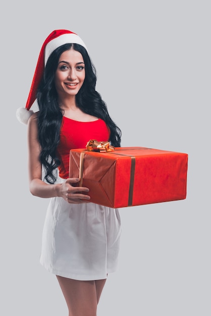 Geschenk für Sie! Attraktive junge Frau mit Weihnachtsmütze, die rote Geschenkbox hält und lächelt, während sie vor grauem Hintergrund steht