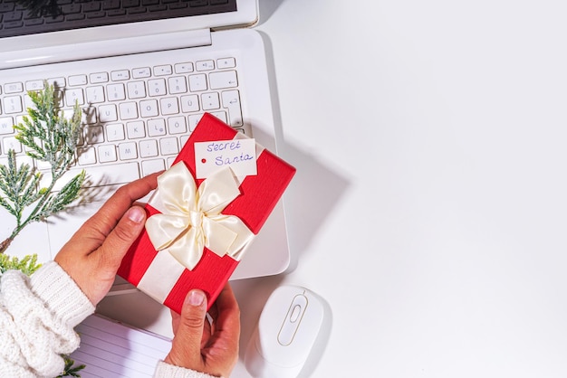 Geschenk vom geheimen Weihnachtsmann auf dem Tisch am Arbeitsplatz. Büro-Weihnachts- und Neujahrsfeierspiel. Bürotisch-Oberflächen-Notizbuch mit festlicher Geschenkbox, weißem Hintergrund, flacher Kopierraum