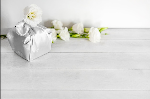 Geschenkbox eingewickelt in Seidenstoff in Furoshiki-Technik weiße Blumen Eustoma heller Holzhintergrund mit Kopierraum Zero Waste Life Concept