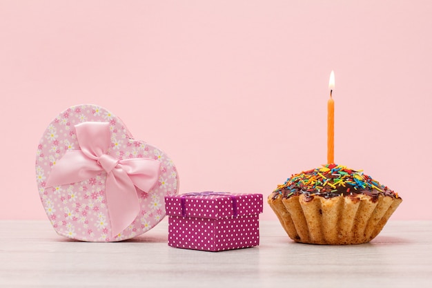 Geschenkbox in Form eines Herzens und eines kleinen, leckeren Geburtstagsmuffins mit Schokoladenglasur und Karamell, dekoriert mit brennender festlicher Kerze auf Holz- und rosafarbenem Hintergrund. Alles Gute zum Geburtstag Konzept.