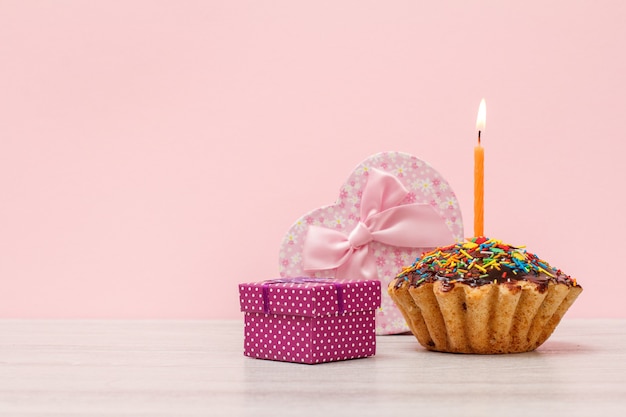 Geschenkbox in Form eines Herzens und eines kleinen, leckeren Geburtstagsmuffins mit Schokoladenglasur und Karamell, dekoriert mit brennender festlicher Kerze auf Holz- und rosafarbenem Hintergrund. Alles Gute zum Geburtstag Konzept.