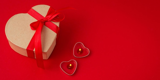 Geschenkbox in Herzform mit Schleife und zwei Kerzen auf rotem Hintergrund Valentinstag