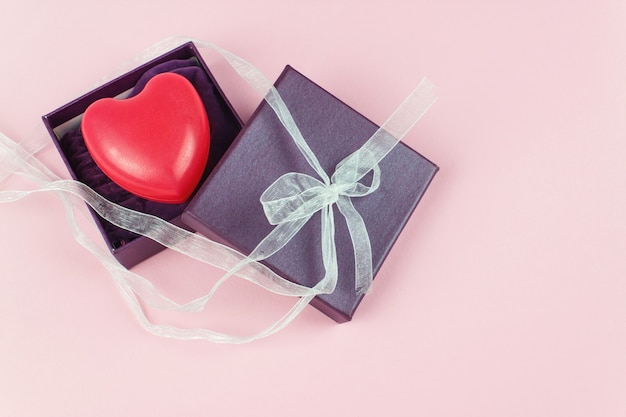 Geschenkbox in lila Farbe mit rotem Herzen auf rosa Hintergrund. Valentinsgruß-Konzept.