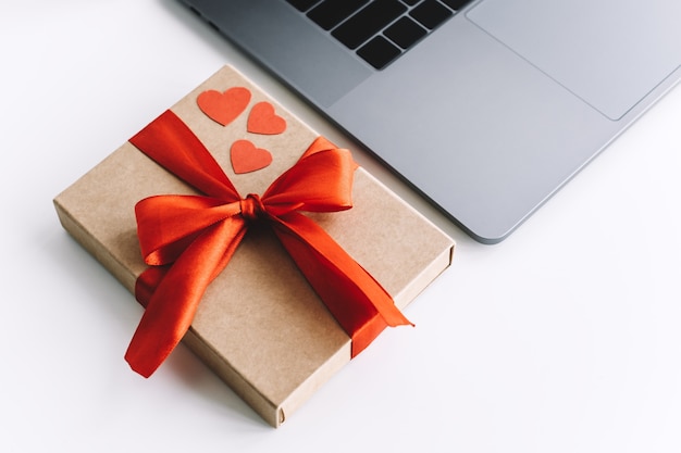 Geschenkbox mit rotem Band und Herzen auf dem Tisch nahe Laptop. Valentinstag Überraschungskonzept