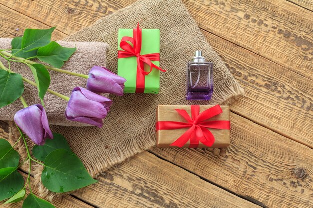 Geschenkboxen, eine Flasche Parfüm und lila Tulpenblumen auf Sackleinen und alten Holzbrettern. Ansicht von oben.