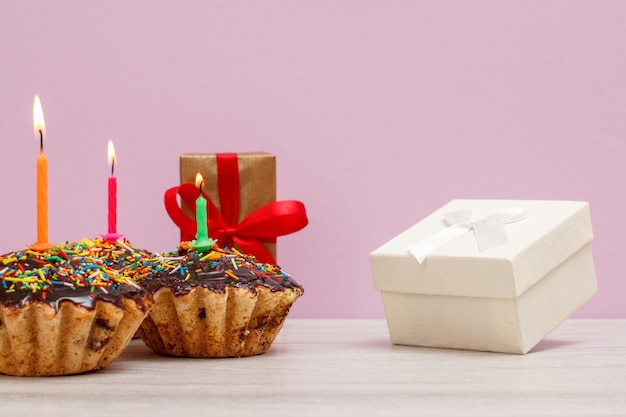 Geschenkboxen und drei leckere Geburtstagsmuffins mit Schokoladenglasur und Karamell, dekoriert mit brennenden festlichen Kerzen auf rosafarbenem Hintergrund. Alles Gute zum Geburtstag minimales Konzept.