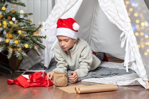 Geschenkverpackung Ein Kind in einer roten Weihnachtsmütze verpackt Weihnachtsgeschenküberraschungen in Kraftpapier mit roten Bändern eines Seils