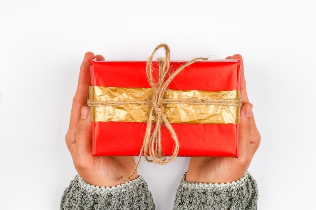 Geschenkverpackung in Rot und Gold in den Händen. Mit einer Schleife auf einem weißen Raum gestrickt. DIY Geschenkverpackung. Frauenhände geben ein verpacktes Geschenk. Weihnachtsgeschenk. Sicht von oben.