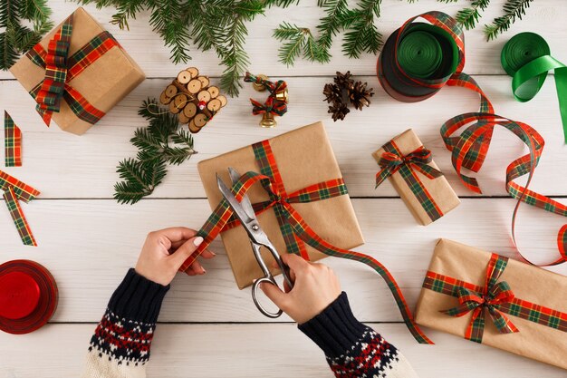 Geschenkverpackungshintergrund. Draufsicht auf weibliche Hände verpacken Weihnachtsgeschenk in Kraftpapier, dekorieren mit Satinband am weißen unordentlichen rustikalen Holztisch. Winterurlaub Konzept.