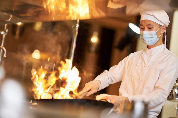 Geschickter Koch mit Gesichtsmaske, der Wok in Flammen hält