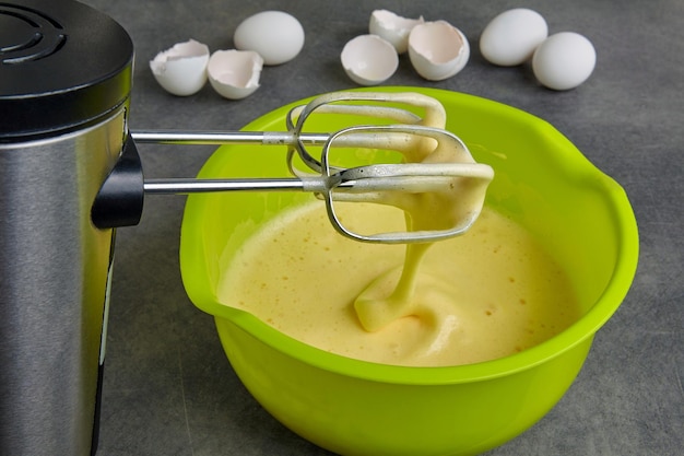 Geschlagene Eier mit Zucker und Mehl in einer grünen Schüssel und einem Mixer vor dem Hintergrund einer Eierschale auf einem dunklen Betontisch