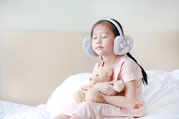 Geschlossene Augen kleines asiatisches Kindermädchen, das Winterohrenschützer trägt und Teddybären beim auf dem Bett zu Hause sitzen umfasst.