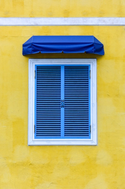 Geschlossenes traditionelles griechisches fenster mit weißem rahmen und blauer gewebemarkise auf gelbem zement