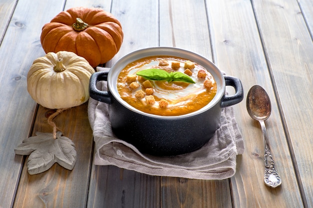 Geschmackvolle Kürbissuppe in der schwarzen keramischen Wanne auf rustikalem Holz. Die Suppe wird mit Creme, Croutons und Basilikumblättern serviert.