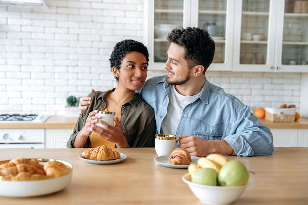 Foto geschmackvolles frühstück glückliches mehrrassiges paar kaukasischer mann und afroamerikanische frau verbringen zeit zusammen zu hause, sie trinken kaffee, essen croissants, plaudern über verschiedene themen, lächeln