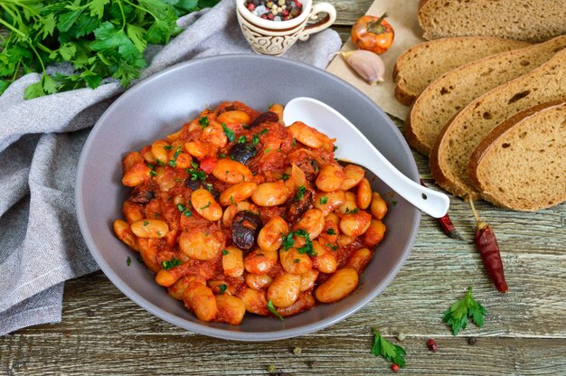 Geschmorte Saubohnen in Tomatensauce mit Kräutern und Gewürzen aus der Nähe, Scheiben Roggenbrot auf dem Holztisch. Fastenmenü. Veganes Gericht.