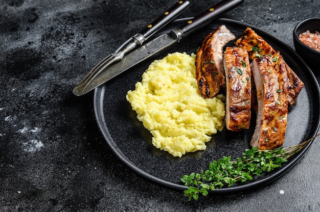 Geschnittene BBQ gegrillte Schweinerippchen auf einem Teller mit Kartoffelpüree. Schwarzer Hintergrund. Ansicht von oben. Platz kopieren.