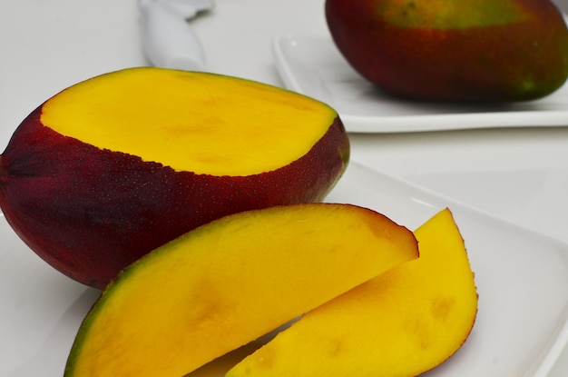 Geschnittene Fruchtmango, innen gelb, Scheiben auf weißem Teller mit Objekten daneben auf weißem Tisch. Mango