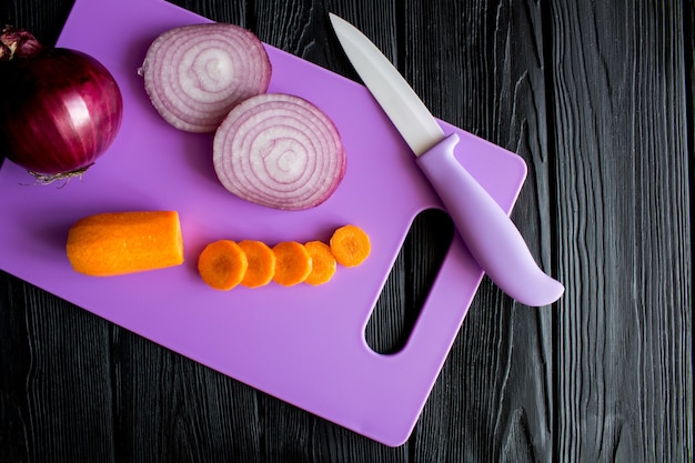 Geschnittene Karotten und Zwiebeln auf dem violetten Schneidebrett auf dem schwarzen hölzernen Hintergrund. Draufsicht.