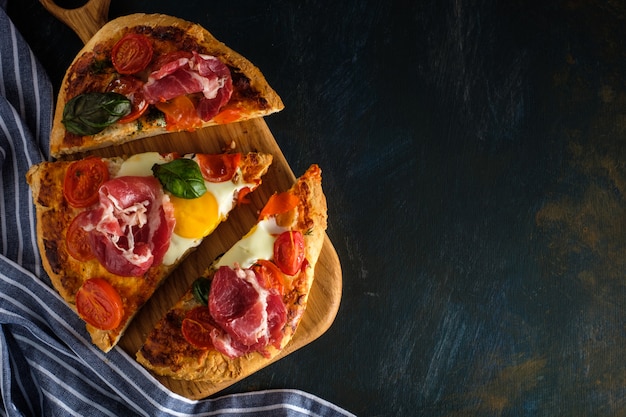 Geschnittene selbst gemachte geschmackvolle Pizza mit Speck, Ei, Käse, Tomaten und Basilikum auf Dunkelheit