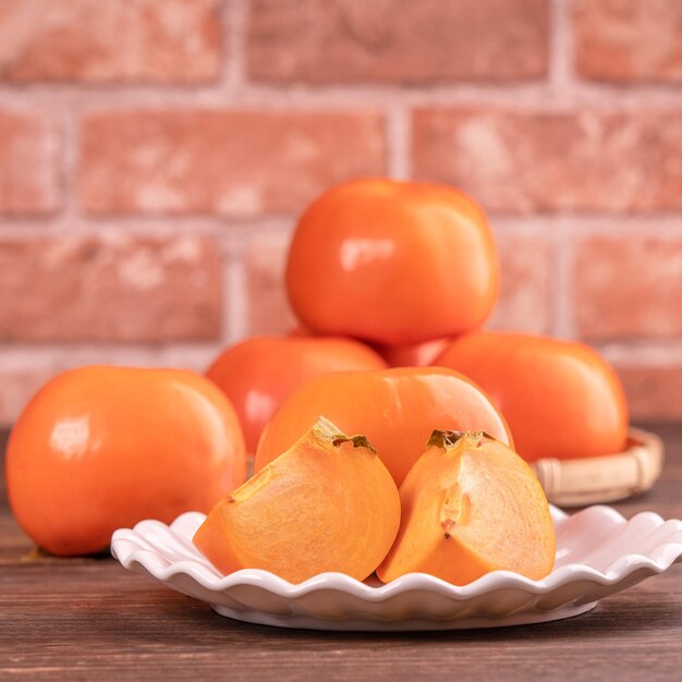 Geschnittene süße Persimmon-Kaki in einem Bambus-Siebkorb auf dunklem Holztisch mit rotem Backsteinmauerhintergrund Chinesisches Neujahrsfrucht-Designkonzept aus nächster Nähe