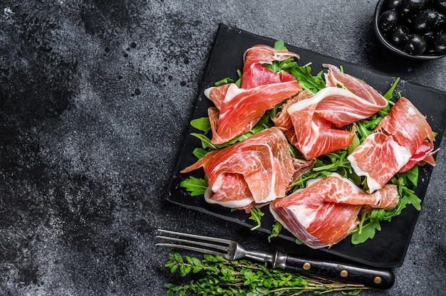 Geschnittenes Jamon Serrano Schweineschinkenfleisch auf einem Marmorbrett. Schwarzer Hintergrund. Ansicht von oben. Platz kopieren.