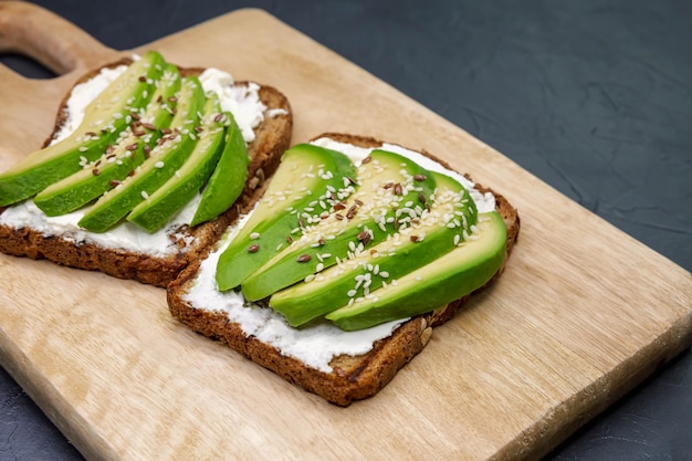 Geschnittenes Sandwich mit Avocado und Hüttenkäse auf Holzbrett auf dunklem Hintergrund